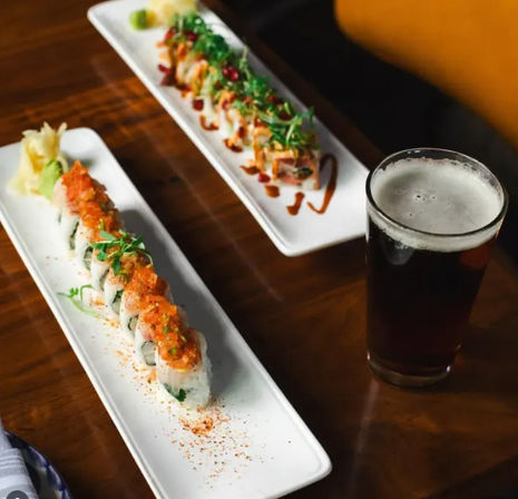 Two long white plates of sushi rolls topped with spicy sauce, microgreens, pickled ginger and wasabi, paired with a pint of dark craft beer on a wooden table.