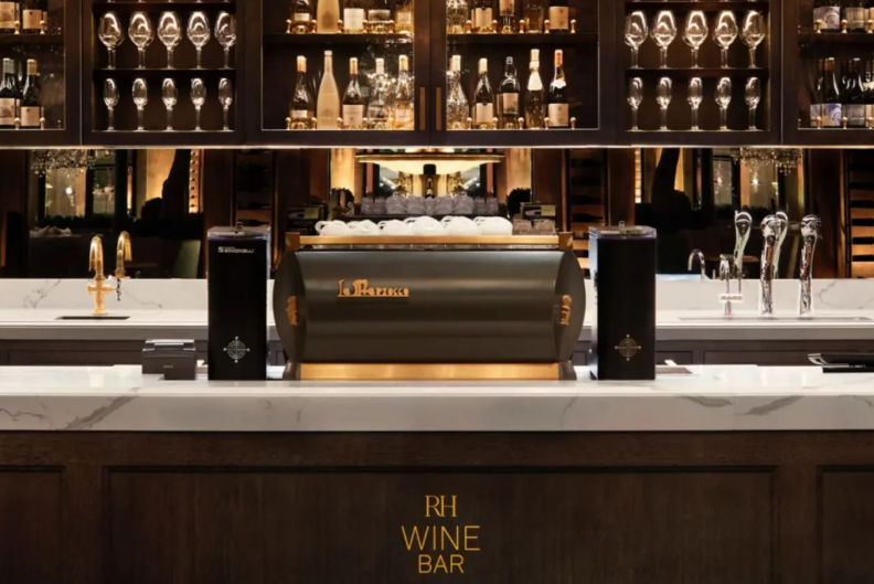 Elegant indoor wine bar with marble countertop, sleek black espresso machine topped with stacked white cups, dark wood bar, backlit shelves of wine bottles and rows of stemware