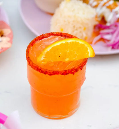 Vibrant orange Mexican‑inspired cocktail/mocktail in a chili-rimmed glass topped with an orange slice, with a blurred plate of rice and pickled onions in the background