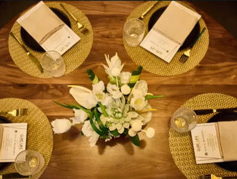 Overhead view of a wooden dining table set for four with woven gold placemats, black plates, gold cutlery, glass tumblers, menus, and a white floral centerpiece with succulents.
