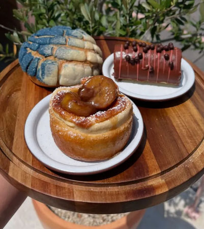 Three artisan pastries on a round wooden serving board: a caramel-glazed rolled pastry topped with caramelized banana, a blue-and-white sugar-topped concha bun, and a chocolate-covered pastry with chocolate pearls in an outdoor setting.