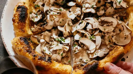 Close-up of gourmet mushroom pizza topped with thinly sliced cremini mushrooms and chives, creamy sauce and a charred golden crust as a hand pulls a slice from a white plate