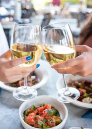 Two hands clinking glasses of white wine above bowls of fresh salad on a marble table at an outdoor café, cheerful al fresco dining scene
