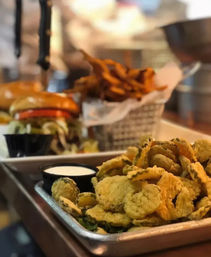 Crispy fried pickle chips with ranch dipping sauce on a metal tray, blurred burger and basket of fries in the background at a casual American restaurant