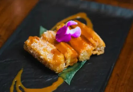 Crispy golden fried dessert sticks drizzled with caramel sauce, garnished with a purple edible orchid on a green leaf, served on a black slate plate.