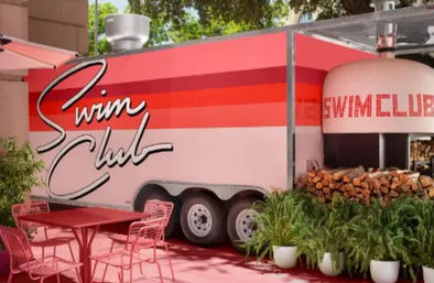 Retro pink-and-red striped food trailer with cursive signage beside a dome wood-fired oven, stacked firewood, red metal patio table and chairs and potted ferns in a sunny outdoor courtyard.
