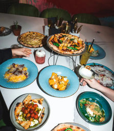 Overhead shot of a lively restaurant table with Neapolitan-style pizzas, pasta and small plates on blue dishes, plus colorful cocktails and hands reaching in — group dining vibe.