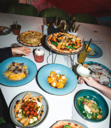 Overhead shot of a lively restaurant table with Neapolitan-style pizzas, pasta and small plates on blue dishes, plus colorful cocktails and hands reaching in — group dining vibe.