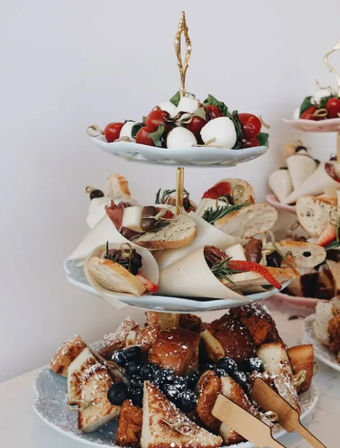 Three-tier ceramic serving stand with caprese skewers on top, assorted crostini with prosciutto and olives on the middle tier, and sweet French toast bites with blueberries and powdered sugar on the bottom — festive brunch or party appetizers.