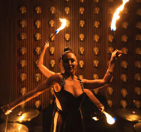 Fire performer in a sleek black outfit twirling flaming torches against a wall of gold skulls in a dim, club-like lounge with percussion cymbals