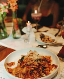 Cozy candlelit Italian restaurant table with a white bowl of rustic pappardelle ragù topped with grated cheese, a glass of red wine, blurred diners and a floral centerpiece