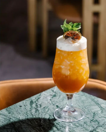 Bright orange tropical cocktail in a stemmed glass with crushed ice, frothy top, dried citrus wheel and mint garnish, sitting on a green marble tabletop.