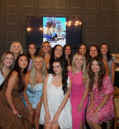 Smiling group photo of young women in colorful dresses at an indoor celebration or party, posing in front of a wall-mounted screen and dark paneled wall