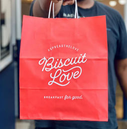 Bright red breakfast takeout bag with white script logo and tagline, held out toward the camera by a person in a dark shirt.