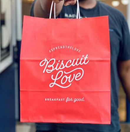 Bright red breakfast takeout bag with white script logo and tagline, held out toward the camera by a person in a dark shirt.