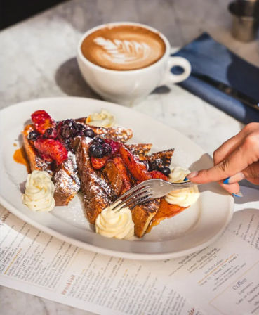 Berry-topped French toast with powdered sugar, whipped-cream rosettes and syrup, a fork reaching in, served with a latte on a marble cafe table — bright brunch scene.
