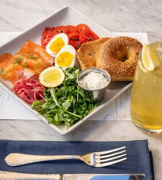Colorful cafe brunch plate with sesame bagel halves, smoked salmon, soft‑boiled eggs, arugula, sliced tomatoes, a cup of cream cheese and a glass of lemon iced tea on a marble table