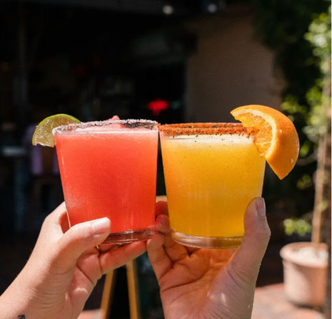 Two frozen margaritas raised on a sunny outdoor patio — a pink lime-salt cocktail with a lime wedge and a yellow-orange chili-rimmed cocktail with an orange slice.