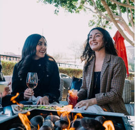 Two friends laughing over wine and a cocktail at a sunny outdoor patio table with appetizers and a fire pit in the foreground.