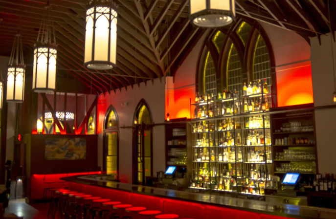 Dimly lit bar interior in a converted church with a vaulted wooden ceiling, arched stained-glass windows, towering backlit liquor shelves, and a long counter glowing with red accent lighting