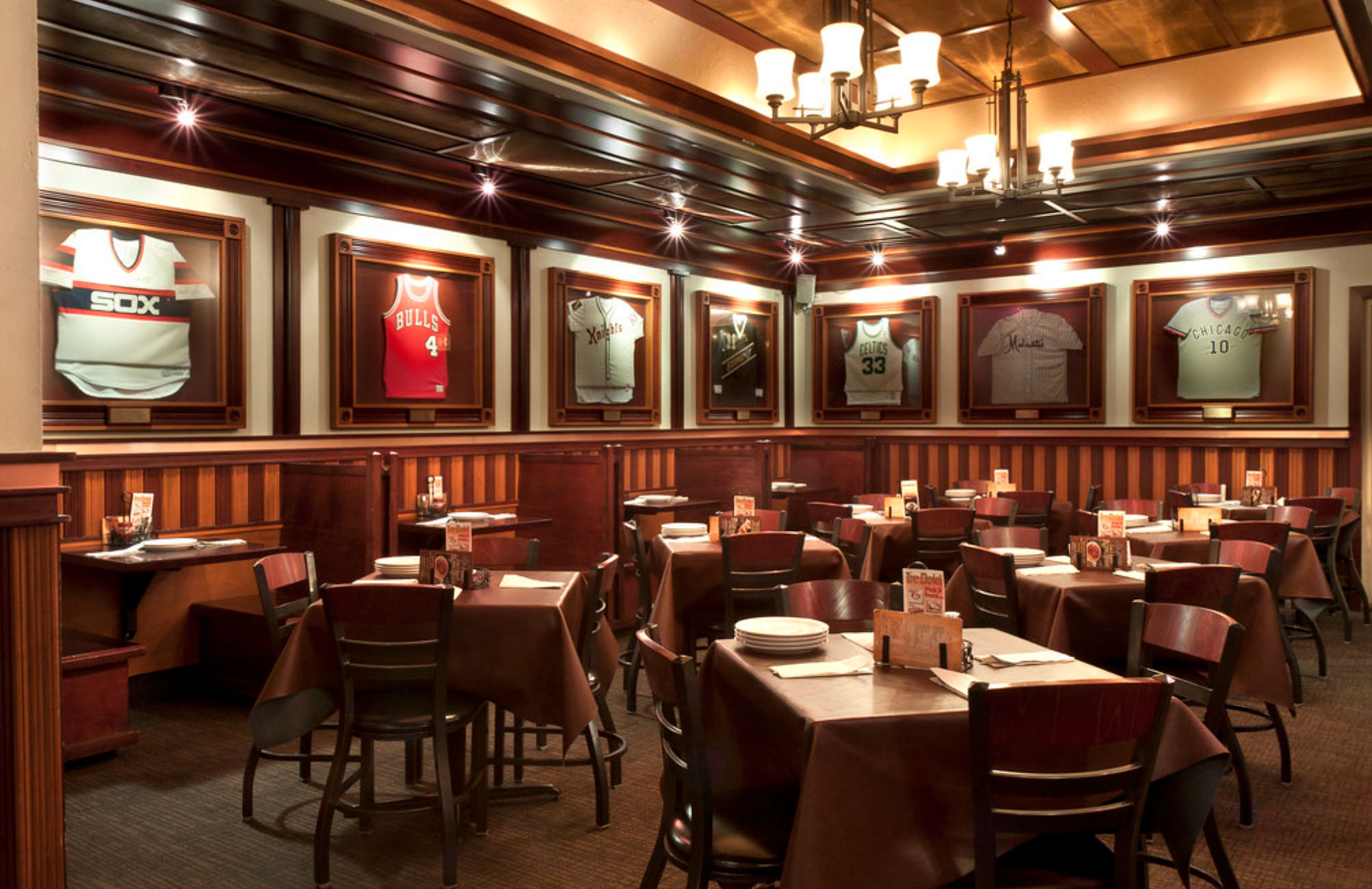 Cozy sports-themed restaurant interior with wood-paneled walls, chandeliers, brown-tablecloth dining tables and framed vintage sports jerseys on display.