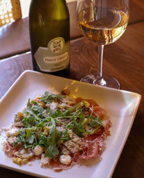 Savory Italian-style appetizer of thin prosciutto topped with arugula, shaved cheese, crunchy crostini and creamy dollops on a white plate, served with a glass and bottle of Pinot Grigio on a wooden table