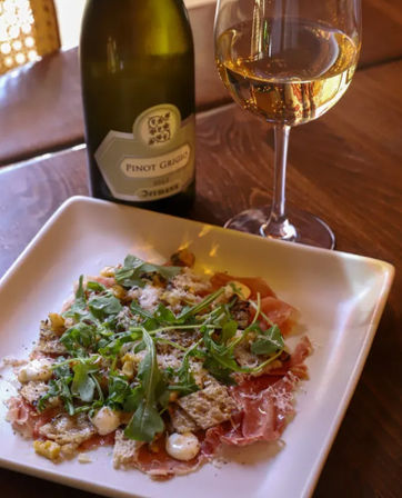 Savory Italian-style appetizer of thin prosciutto topped with arugula, shaved cheese, crunchy crostini and creamy dollops on a white plate, served with a glass and bottle of Pinot Grigio on a wooden table