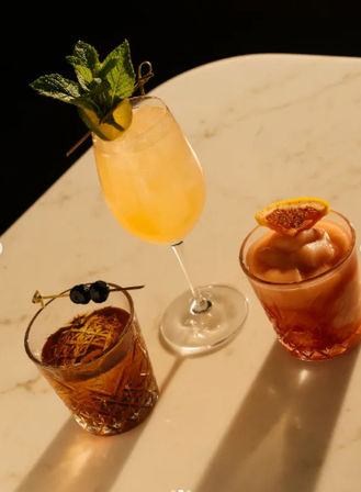 Three craft cocktails on a marble bar top: a tall peachy spritz garnished with mint and cucumber, a crystal lowball with dark cherry skewers and citrus zest, and a frozen pink drink topped with a candied orange slice.