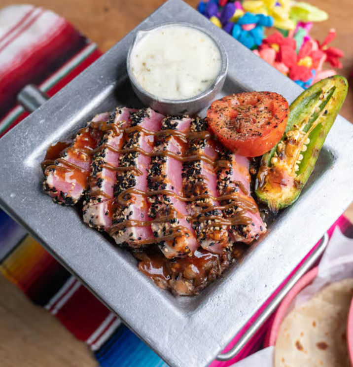 Seared sesame-crusted tuna slices drizzled with glaze on a square metal plate, served with a charred jalapeño, grilled tomato and creamy dipping sauce over a colorful Mexican serape backdrop.