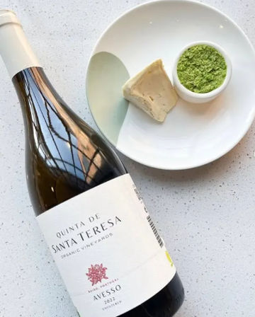 Bottle of white wine on its side next to a small plate with a wedge of cheese and a ramekin of bright green herb pesto on a speckled white countertop — playful wine-and-cheese pairing