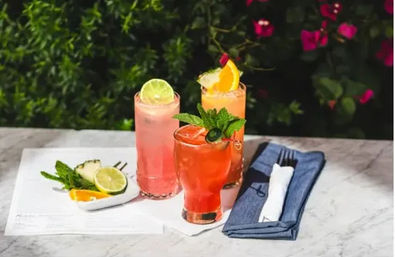 Three colorful summer cocktails on a marble outdoor patio table—pink lime soda, orange citrus spritz, and red fruit cocktail garnished with mint and a green slice—served with citrus wedges and a folded blue napkin, lush garden with pink flowers in the background.
