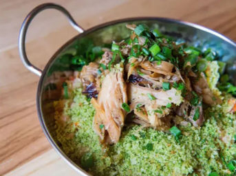 Shredded pulled chicken piled on bright green herbed couscous, topped with chopped scallions in a small metal serving bowl on a wooden table.