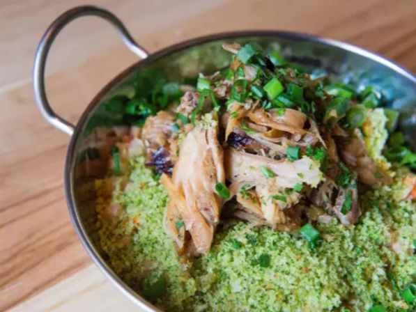 Shredded pulled chicken piled on bright green herbed couscous, topped with chopped scallions in a small metal serving bowl on a wooden table.