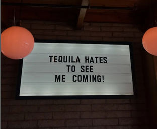 Backlit marquee sign on a brick wall in a bar interior reading "TEQUILA HATES TO SEE ME COMING!" flanked by two round pendant lamps — humorous cocktail-themed decor.