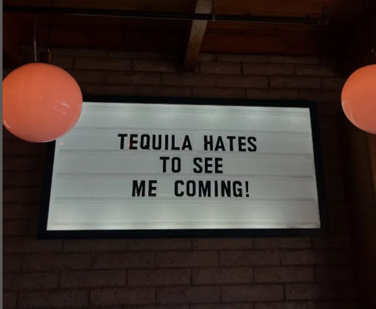 Backlit marquee sign on a brick wall in a bar interior reading "TEQUILA HATES TO SEE ME COMING!" flanked by two round pendant lamps — humorous cocktail-themed decor.