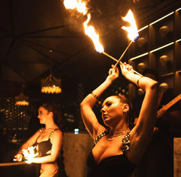 Two fire dancers twirling flaming torches inside a dim, upscale nightclub lounge with amber chandeliers and moody stage lighting, indoor fire performance entertainment.