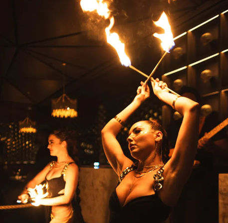 Two fire dancers twirling flaming torches inside a dim, upscale nightclub lounge with amber chandeliers and moody stage lighting, indoor fire performance entertainment.
