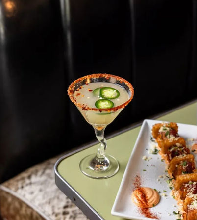 Zesty jalapeño margarita in a twisted-stem martini glass with chili-salt rim and fresh jalapeño slices, served next to a white plate of crispy fried bites and creamy paprika dipping sauce on a retro green tabletop.