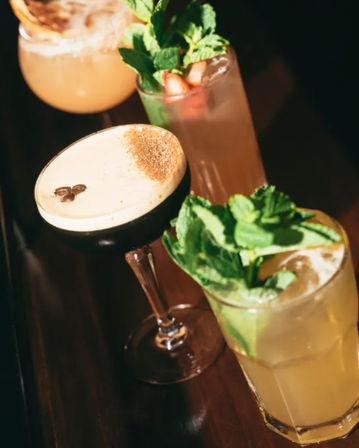 Close-up of vibrant cocktails on a dark bar counter: an espresso martini topped with coffee beans, a mojito-style lime drink with fresh mint, and two fruit-garnished mixed cocktails—stylish cocktail bar vibes.