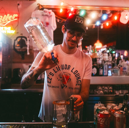 Bartender in a cap and glasses pouring clear liquor into a jigger while mixing a cocktail behind a cozy neon-lit neighborhood bar, with colorful string lights, bottles on shelves, and soda cans on the counter.