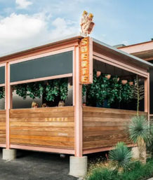 Covered outdoor dining patio with pastel pink metal frame and wooden slat railing, hanging greenery, warm illuminated vertical sign and desert-style plants nearby.