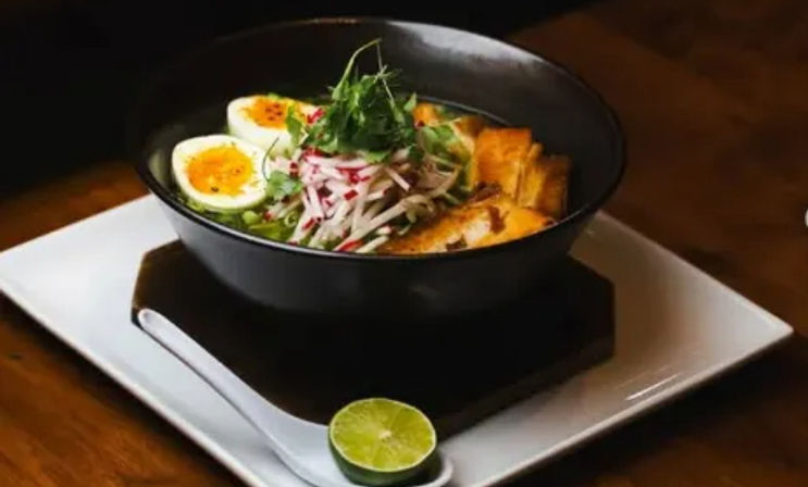 Black bowl of ramen-style noodle soup with halved soft-boiled eggs, shredded radish, fresh greens and seared tofu, served on a white square plate with a lime half on a ceramic spoon on a wooden table