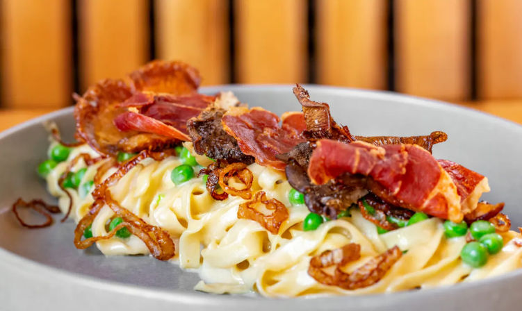 Close-up of restaurant-style creamy fettuccine pasta with green peas, fried shallots and crispy cured-meat ribbons on a gray plate.