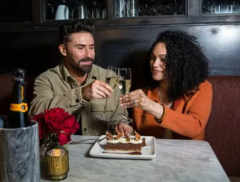 Smiling couple toasting with champagne flutes over dessert at a candlelit restaurant table with red roses and a chilled bottle