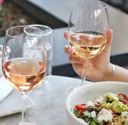 Two glasses of rosé wine on a marble table, one held by a hand, next to a fresh avocado and tomato salad in a white bowl at an outdoor cafe