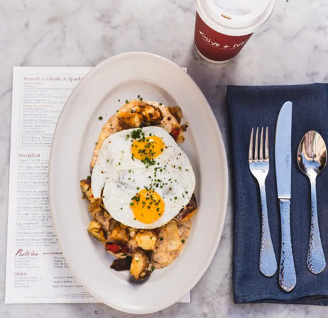 Top-down view of a cafe brunch: oval white plate with two sunny-side-up eggs over roasted potato hash and toast, menu peeking underneath, takeaway coffee cup and fork-knife-spoon on a navy napkin on a marble table.