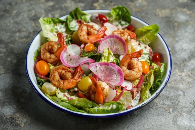 Grilled shrimp salad with mixed greens, sliced pink radish, yellow and red cherry tomatoes, pickled onions and creamy dressing served in an enamel bowl on a stone countertop
