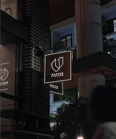 Glowing square restaurant sign with minimalist logo on a brick facade at night, modern Italian dining vibe on an urban street
