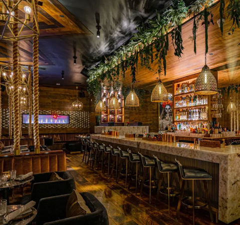 Wood‑paneled upscale cocktail bar with long marble counter, leather bar stools, woven pendant lights, hanging greenery and intimate lounge seating
