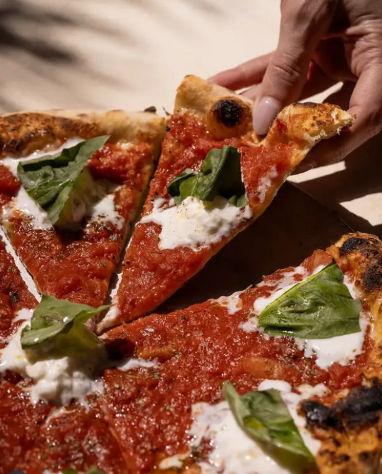 Close-up of a hand grabbing a slice of wood-fired Neapolitan-style margherita pizza with bright tomato sauce, creamy mozzarella dollops, fresh basil leaves and charred thin crust.
